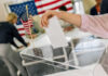 Hand placing ballot in box with American flag nearby