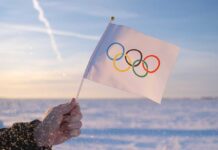 A hand holding an Olympic flag against a snowy landscape during sunset