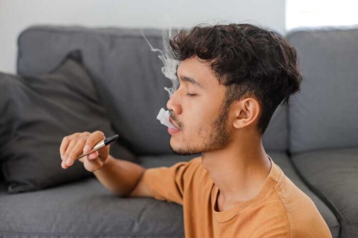 Young man sitting on a couch, vaping with smoke around him