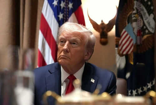 trump-oval-room2595875411 Man in suit sitting with flag background and eagle decoration