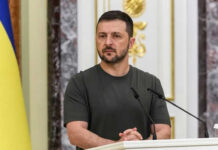 Man speaking at podium with microphones, Ukrainian flag behind.