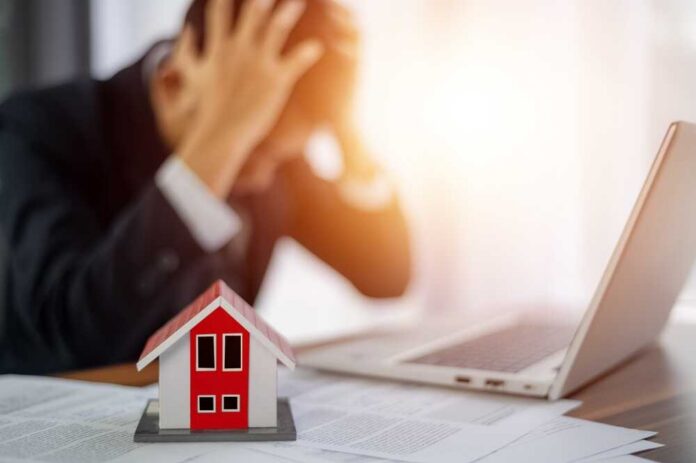A businessman with his head in his hands, stressed over paperwork and a model house on the table