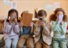 Four children sitting together, each focused on their electronic devices