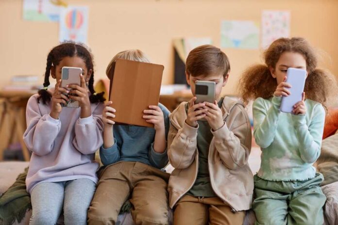 Four children sitting together, each focused on their electronic devices