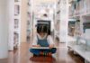 A young girl sitting on the floor of a library, reading a book