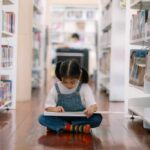 A young girl sitting on the floor of a library, reading a book
