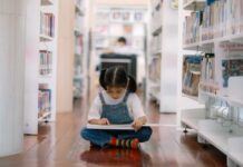 A young girl sitting on the floor of a library, reading a book