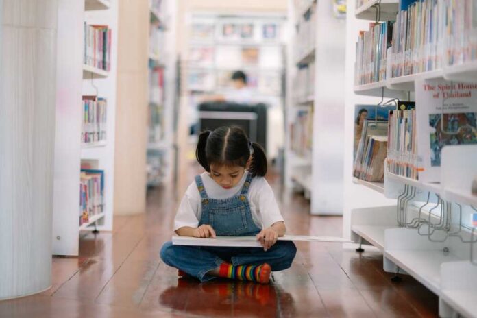 A young girl sitting on the floor of a library, reading a book