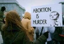 DOJ’s Secret Abortion Loophole Exposed Person holding a sign that reads 'Abortion is Murder' at a protest