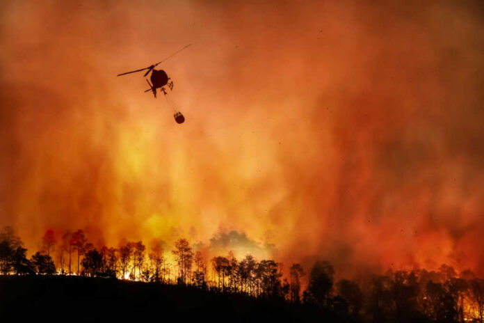 Helicopter fighting wildfire over burning forest at dusk