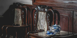 A wooden table with a blue and white teapot and cup next to ornate wooden chairs