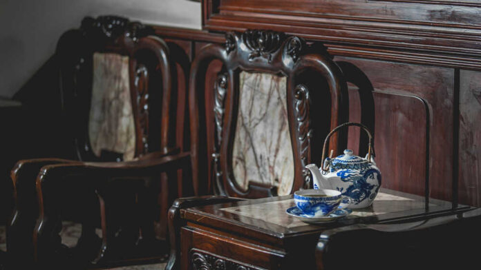 A wooden table with a blue and white teapot and cup next to ornate wooden chairs