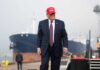 A man in a suit wearing a red cap stands near a ship at a harbor