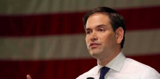 man speaking with microphone in front of an American flag