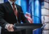 Whisked Out Before Police Knocked – Airlift Sparks Cover-Up man in suit at podium with microphones