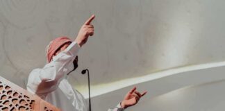Speaker in traditional attire gesturing from a carved wooden podium
