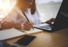 person writing in notebook while using a laptop