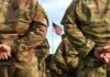 Curfew Slams Troops After Hotel Death Soldiers in camouflage stand with hands behind backs facing a U.S. flag