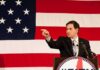 Speaker in suit pointing during speech before large American flag